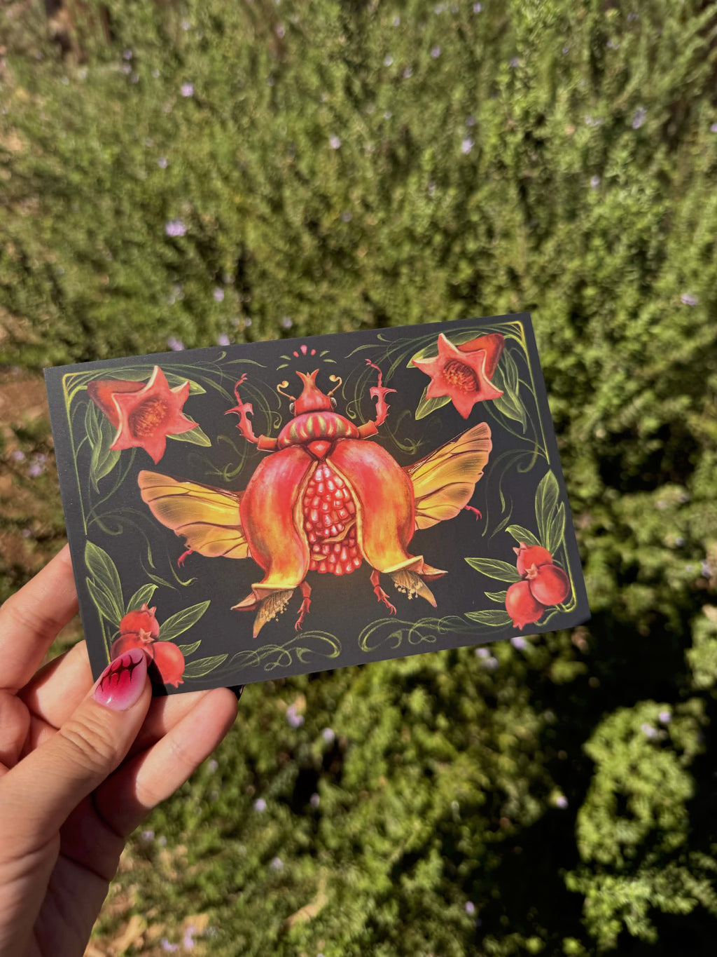 A hand holds a rectangular art print or card featuring a colorful beetle illustration with red, orange, and yellow tones on a dark background, set against a blurred green outdoor backdrop.
